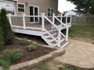 Tan paver patio adjoining a backyard deck with white railings and black balusters