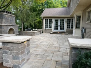 Gray paver patio with a stone outdoor fireplace and low retaining walls beside a home